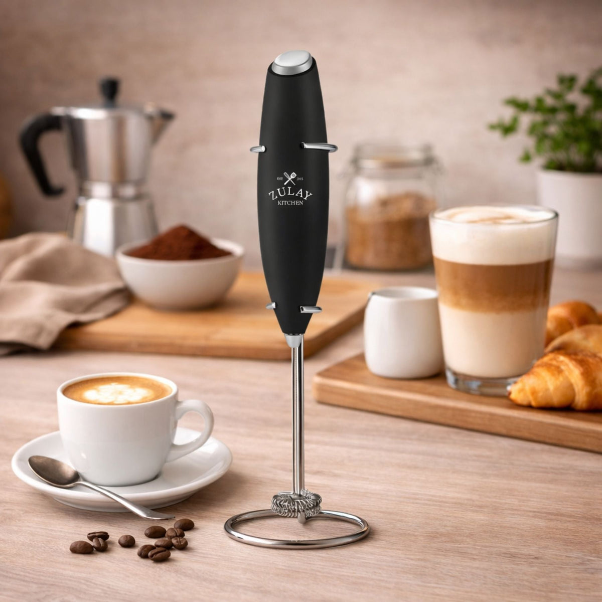 Handheld coffee frother on a kitchen counter with coffee and croissants in the background