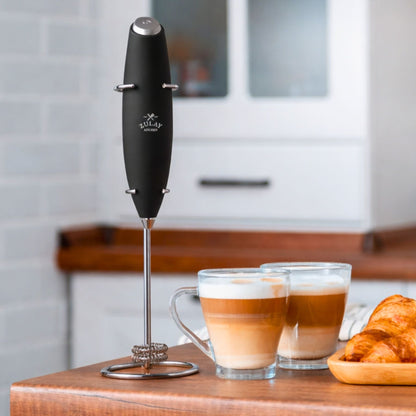 Black milk frother with a stand on a kitchen counter next to two coffee cups and a croissant.